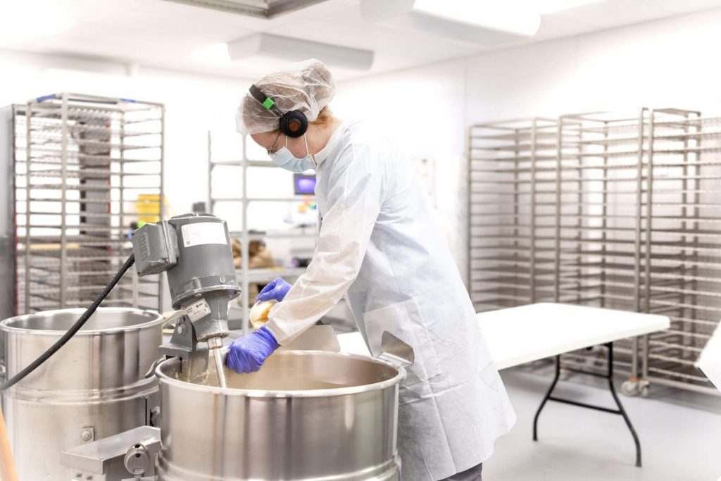 A person wearing a lab coat and protective hair net and mask mixing something in a tank